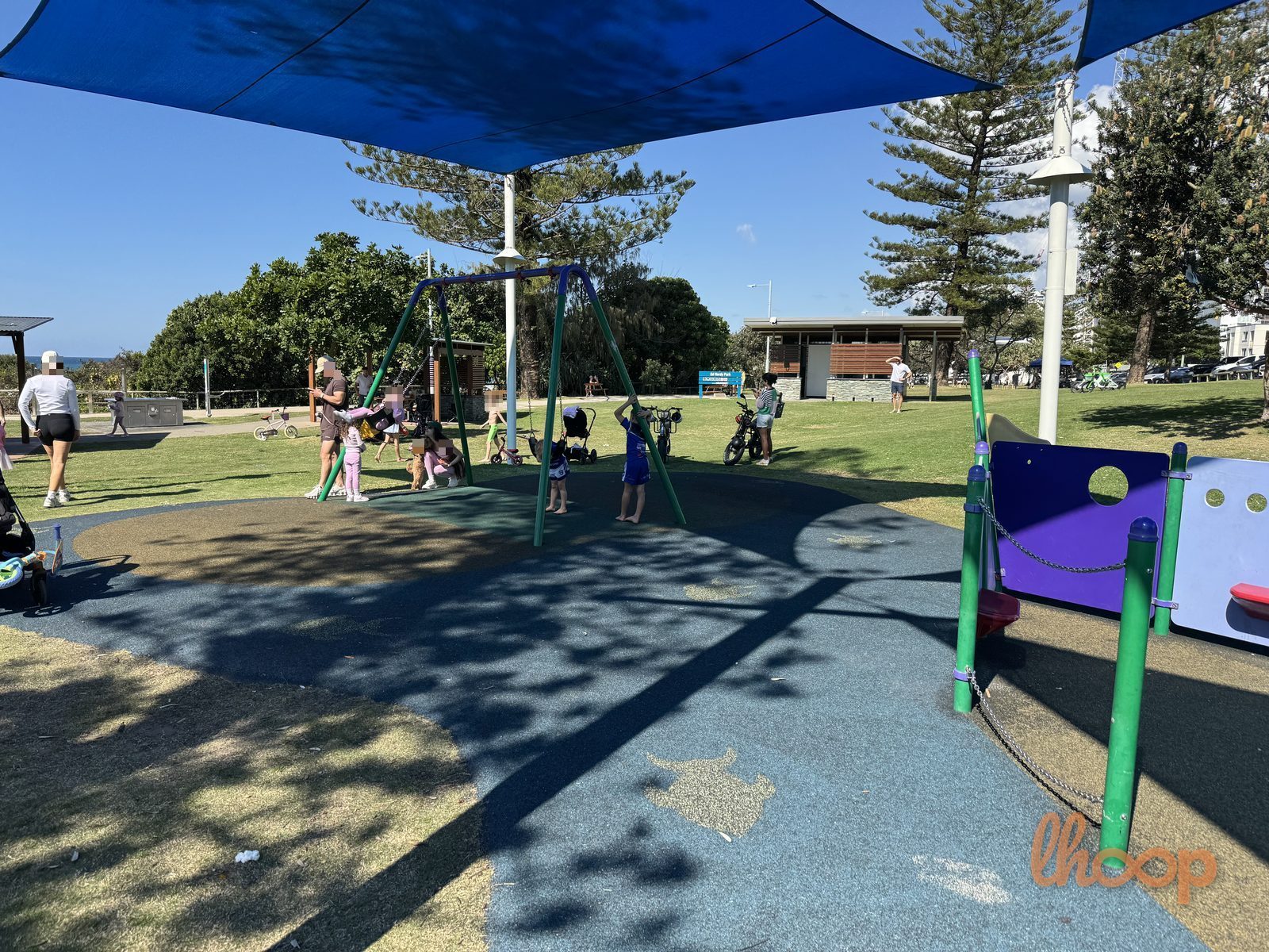 The swing set under a shade sail, with the lawn, a toilet block and the beachfront path beyond.