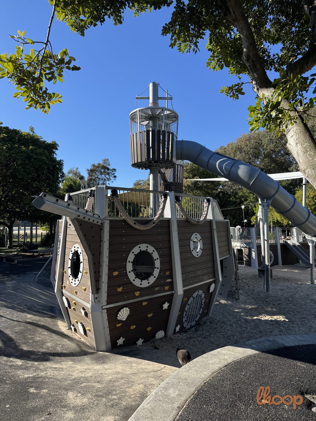 The bow of the timber pirate ship with portholes, climbing holds and rope rails, the crow's nest tower and tunnel slide rising behind it.