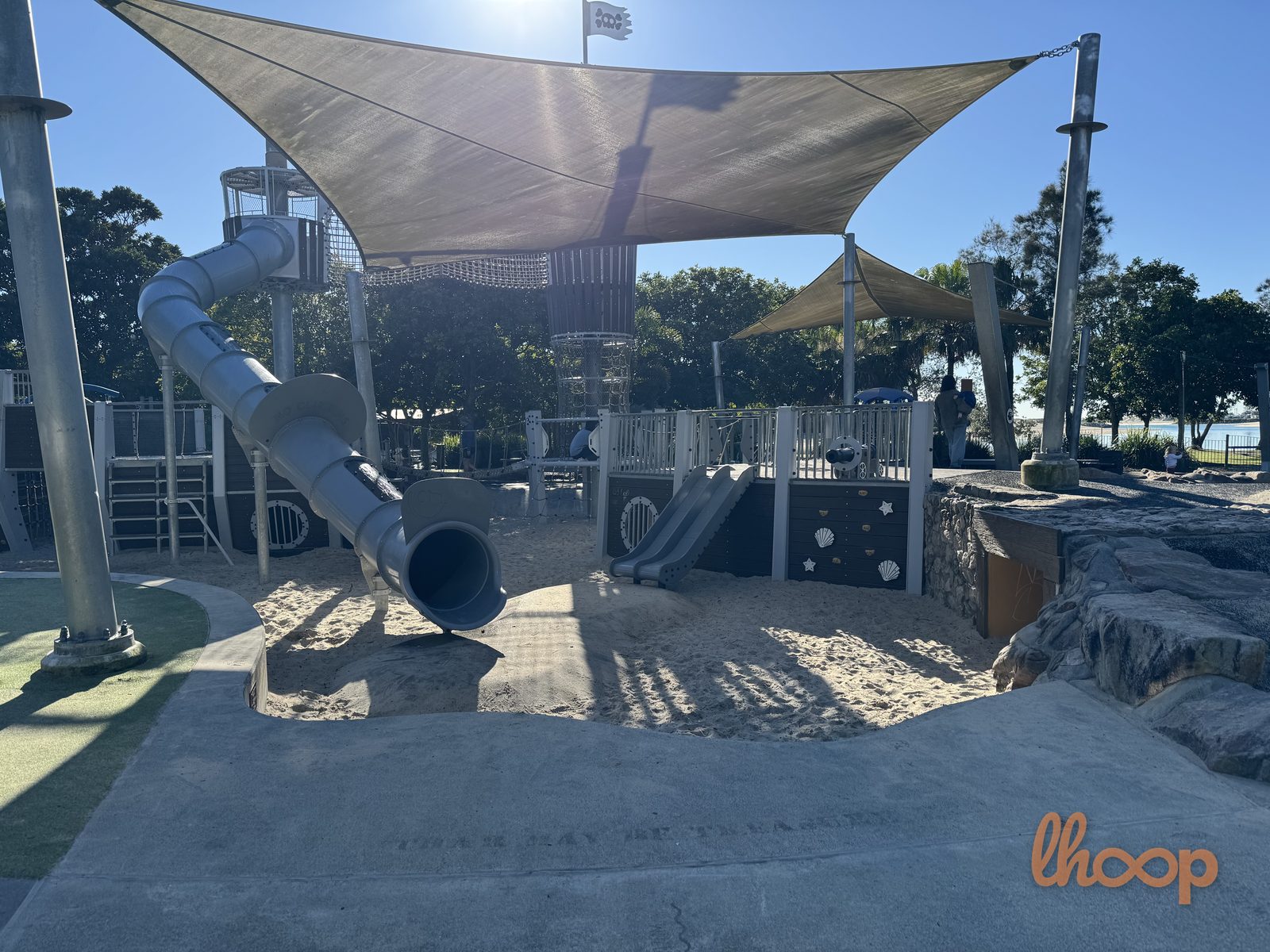 The grey tunnel slide landing on the sand under a big shade sail, beside a smaller slide and shell-patterned panels for younger children.