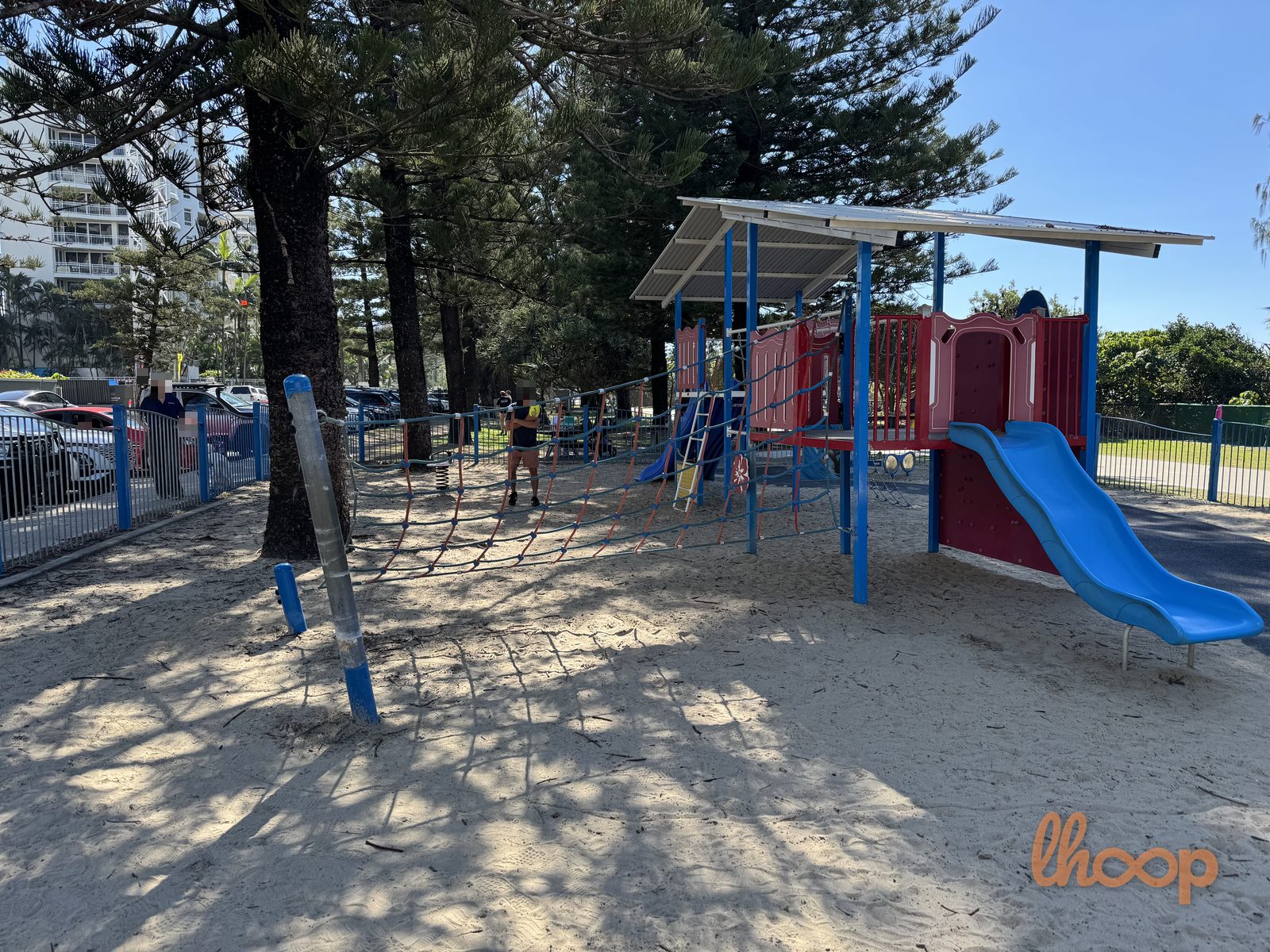 The main play unit under its shade roof, with a blue wave slide, a climbing net and deep sand underfoot.