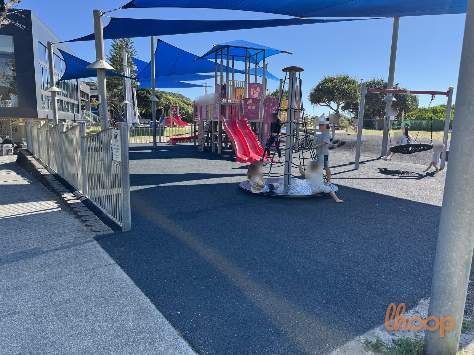 Wide view of the fenced play area with the rope merry-go-round, red slides, nest swing and shade sails on rubber softfall.